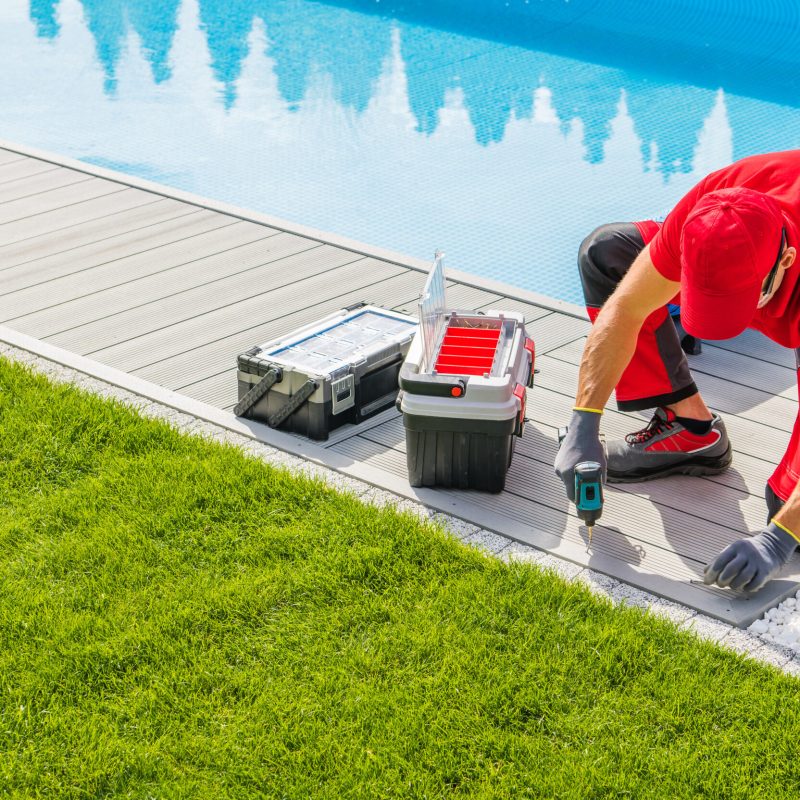 Caucasian Professional Worker Doing a Pool Deck Installation Around Outdoor Swimming Pool in His Client's Backyard. Toolboxes Next to Him.
